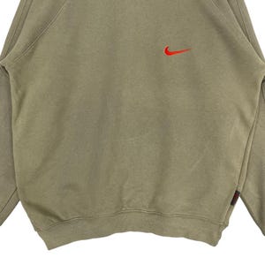 May include: A tan-colored sweatshirt with a red Nike swoosh logo on the upper chest. The long-sleeved top has a crew neck and a small red tag on the lower left side. The fabric appears to be a soft, comfortable material.
