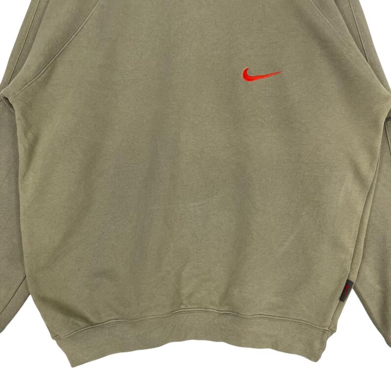 May include: A tan-colored sweatshirt with a red Nike swoosh logo on the upper chest. The long-sleeved top has a crew neck and a small red tag on the lower left side. The fabric appears to be a soft, comfortable material.