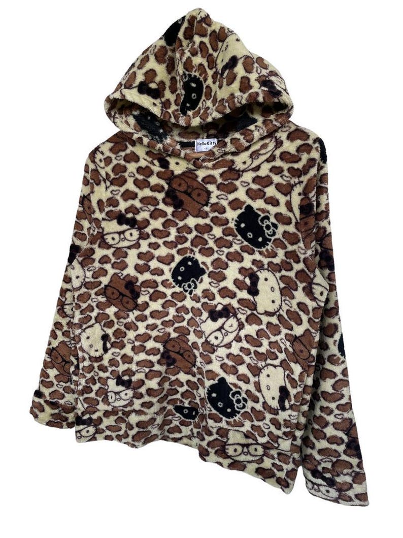 Może przedstawiać: Brązowa i beżowa bluza z kapturem z nadrukiem w cętki. Bluza posiada wiele wzor&oacute;w postaci Hello Kitty w kolorze brązowym, czarnym i białym. Kaptur jest podszyty ciemniejszą brązową tkaniną.