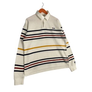 May include: A long-sleeve off-white rugby shirt featuring a collar and a button placket. The shirt is decorated with horizontal stripes in navy blue, dark red, and mustard yellow. The Fila logo is embroidered in navy blue on the left chest area.