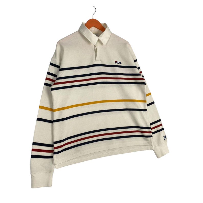May include: A long-sleeve off-white rugby shirt featuring a collar and a button placket. The shirt is decorated with horizontal stripes in navy blue, dark red, and mustard yellow. The Fila logo is embroidered in navy blue on the left chest area.