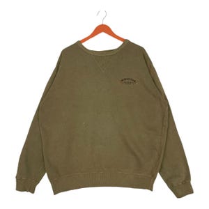 May include: An olive green sweatshirt with a crew neck and long sleeves. The garment has a small embroidered design above the chest. The sweatshirt is suspended from a wooden hanger.