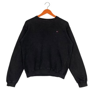 May include: Black crew neck sweatshirt with a small red, white, and blue logo on the chest. The sweatshirt is hanging on a wooden hanger against a white background. The garment has long sleeves and a ribbed neckline, cuffs, and hem.