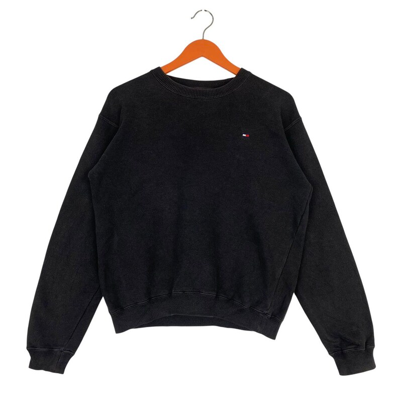 May include: Black crew neck sweatshirt with a small red, white, and blue logo on the chest. The sweatshirt is hanging on a wooden hanger against a white background. The garment has long sleeves and a ribbed neckline, cuffs, and hem.