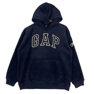 May include: A navy blue fleece hoodie featuring a large white GAP logo across the chest. The garment includes a hood, long sleeves with ribbed cuffs, a front kangaroo pocket, and a small rectangular patch on the left sleeve with white text.