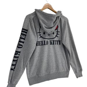 May include: A gray hooded sweatshirt featuring a Hello Kitty graphic on the back and the words "HELLO KITTY" printed on the left sleeve. The Hello Kitty design includes a red bow. The sweatshirt is hanging on a wooden hanger.