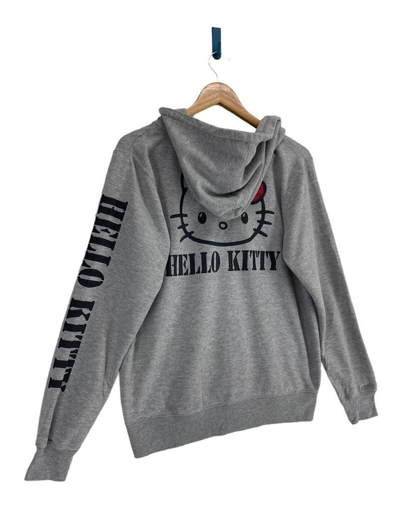 May include: A gray hooded sweatshirt featuring a Hello Kitty graphic on the back and the words "HELLO KITTY" printed on the left sleeve. The Hello Kitty design includes a red bow. The sweatshirt is hanging on a wooden hanger.