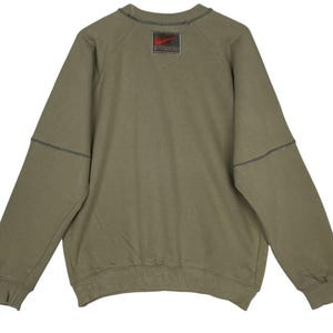 May include: Olive green sweatshirt with a crew neck and long sleeves. Features a stitched design on the sleeves and a red and black logo patch on the upper back. The sweatshirt has a relaxed fit.