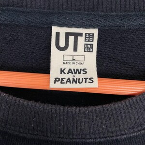 May include: Close-up of a black sweatshirt tag with the white text "UT" and "UNIQLO". Below the "UT" logo is the text "L" and "MADE IN CHINA". The tag also features the text "KAWS x PEANUTS".