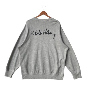 May include: A heather gray sweatshirt with long sleeves, hanging on a wooden hanger. The back of the sweatshirt features the black signature of Keith Haring. The sweatshirt has a crew neck and ribbed cuffs and hem.