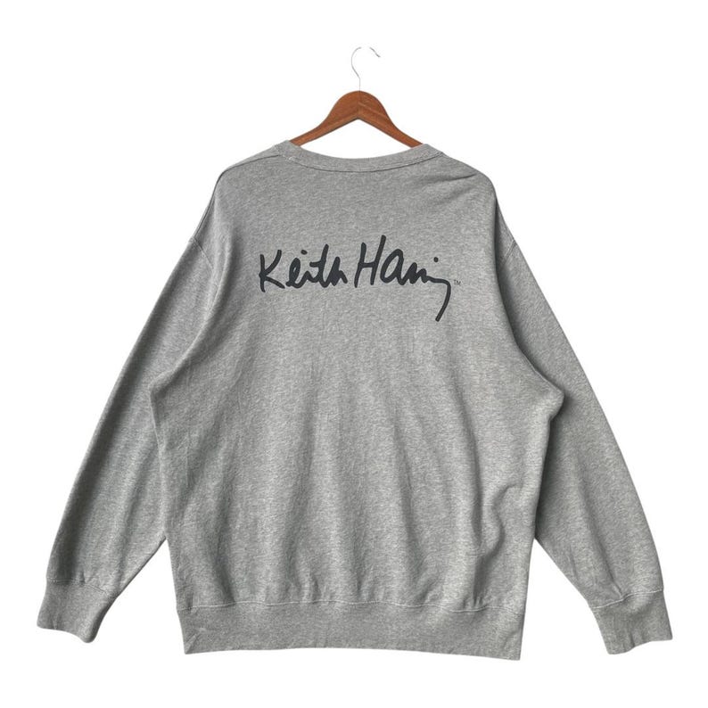May include: A heather gray sweatshirt with long sleeves, hanging on a wooden hanger. The back of the sweatshirt features the black signature of Keith Haring. The sweatshirt has a crew neck and ribbed cuffs and hem.
