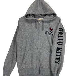 May include: A gray zip-up hoodie featuring the Hello Kitty logo on the chest and the words "Hello Kitty" printed on the sleeve. The hoodie has a hood and a front pocket.