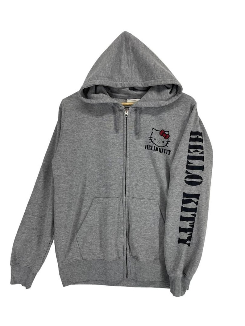 May include: A gray zip-up hoodie featuring the Hello Kitty logo on the chest and the words "Hello Kitty" printed on the sleeve. The hoodie has a hood and a front pocket.
