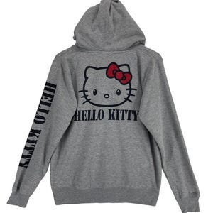 May include: A gray hooded sweatshirt featuring a Hello Kitty graphic on the back with a red bow and the words "HELLO KITTY" in black. The left sleeve also has the words "HELLO KITTY" printed vertically in black.