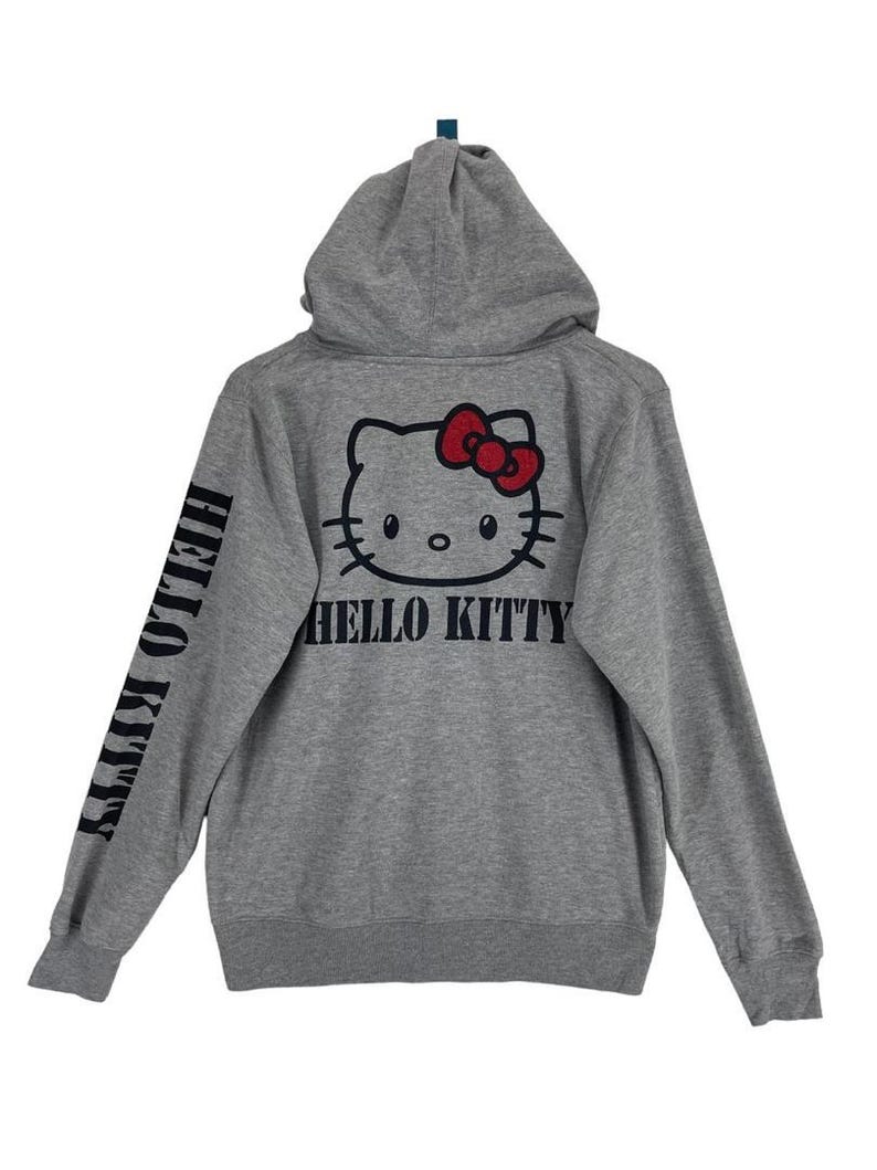 May include: A gray hooded sweatshirt featuring a Hello Kitty graphic on the back with a red bow and the words "HELLO KITTY" in black. The left sleeve also has the words "HELLO KITTY" printed vertically in black.