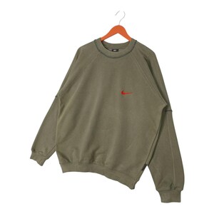 May include: Olive green sweatshirt with a small red Nike logo on the chest. The long-sleeved top has a crew neck and is hanging on a wooden hanger. The stitching is visible in black.