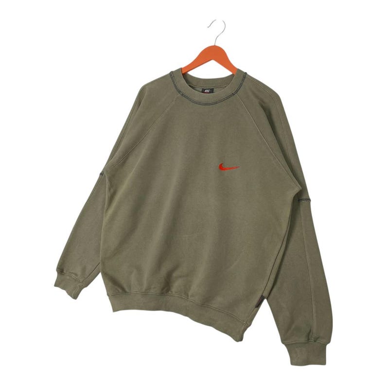 May include: Olive green sweatshirt with a small red Nike logo on the chest. The long-sleeved top has a crew neck and is hanging on a wooden hanger. The stitching is visible in black.