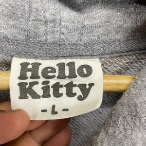 May include: Close-up of a gray hoodie with a tag that reads "Hello Kitty" in a bold, rounded font. The tag also has the size "-L-" printed below the brand name. The hoodie is made of a soft, textured fabric.