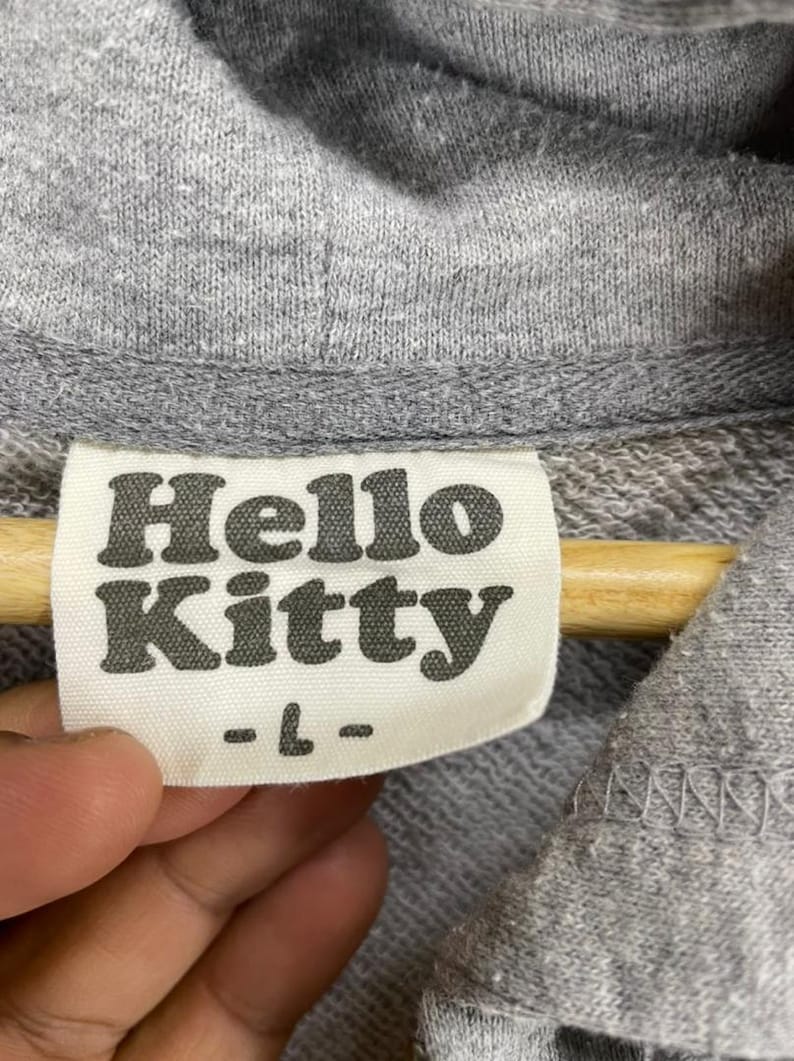 May include: Close-up of a gray hoodie with a tag that reads "Hello Kitty" in a bold, rounded font. The tag also has the size "-L-" printed below the brand name. The hoodie is made of a soft, textured fabric.
