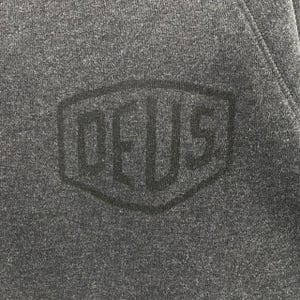 Pode incluir: Close-up de um moletom cinza escuro com uma superf&iacute;cie texturizada. Um logotipo preto em relevo com a palavra "DEUS" est&aacute; centralizado no peito. O logotipo est&aacute; dentro de uma forma hexagonal.