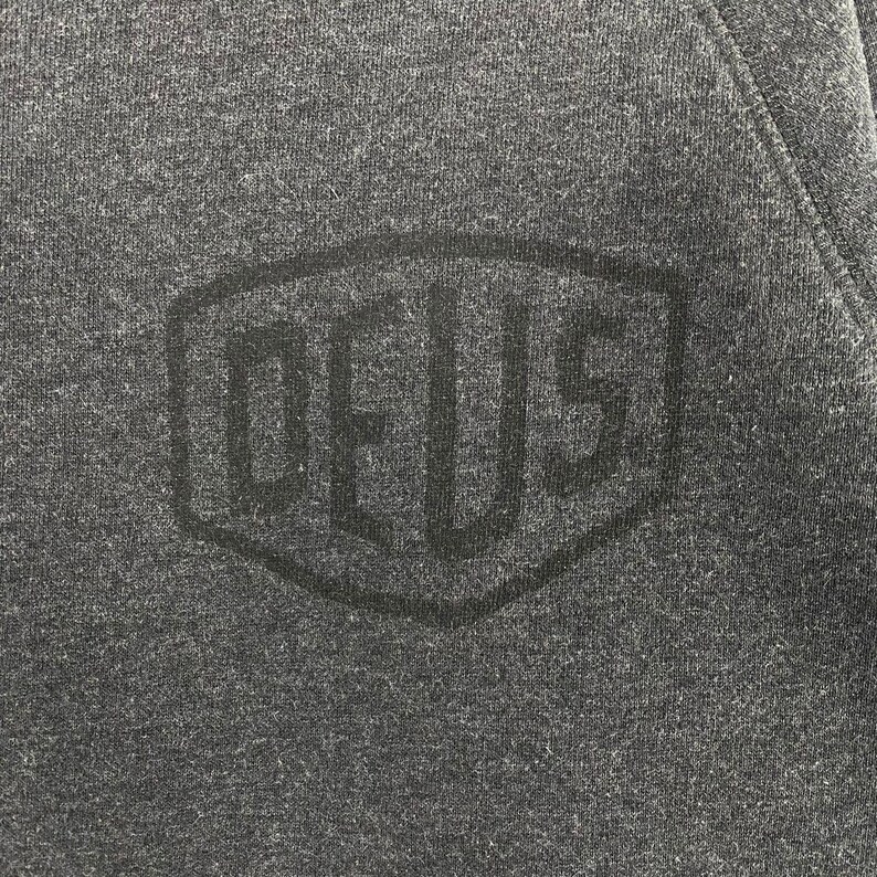 Pode incluir: Close-up de um moletom cinza escuro com uma superf&iacute;cie texturizada. Um logotipo preto em relevo com a palavra "DEUS" est&aacute; centralizado no peito. O logotipo est&aacute; dentro de uma forma hexagonal.