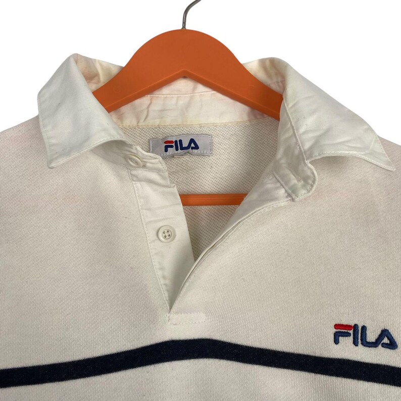 May include: A white long-sleeve polo shirt featuring a navy blue horizontal stripe across the chest. The shirt has a white collar, a button-up placket, and the Fila logo embroidered in navy blue and red on the chest and on the interior neck label.