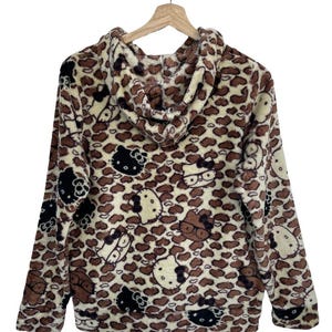 Może przedstawiać: Bluza z kapturem z brązowym i kremowym wzorem w cętki. Bluza posiada wiele wzor&oacute;w postaci Hello Kitty w kolorze czarnym, brązowym i białym. Odzież wisi na drewnianym wieszaku na białym tle.