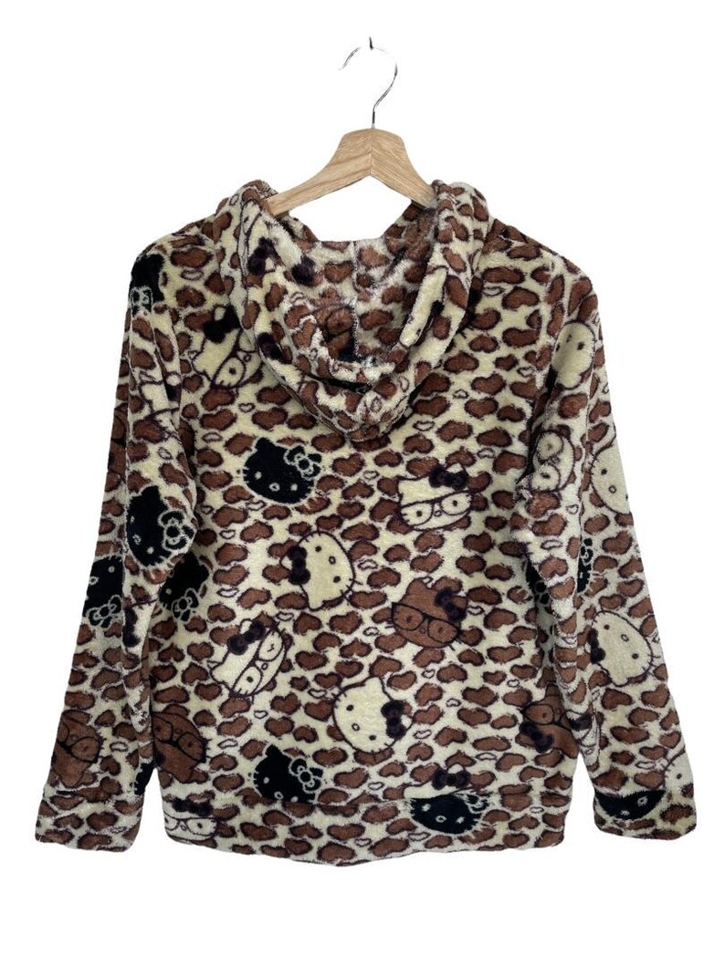 Może przedstawiać: Bluza z kapturem z brązowym i kremowym wzorem w cętki. Bluza posiada wiele wzor&oacute;w postaci Hello Kitty w kolorze czarnym, brązowym i białym. Odzież wisi na drewnianym wieszaku na białym tle.