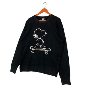 May include: Black crew neck sweatshirt featuring a white graphic of Snoopy skateboarding. The sweatshirt has long sleeves and a ribbed neckline and cuffs. The graphic is a simple line drawing of Snoopy on a skateboard.