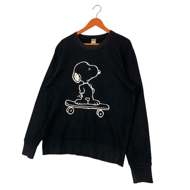 May include: Black crew neck sweatshirt featuring a white graphic of Snoopy skateboarding. The sweatshirt has long sleeves and a ribbed neckline and cuffs. The graphic is a simple line drawing of Snoopy on a skateboard.
