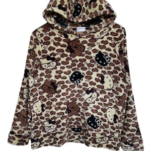 Może przedstawiać: Bluza z kapturem z wzorem w cętki i postaciami Hello Kitty. Bluza wykonana jest z miękkiego, pluszowego materiału z kapturem ze sznurkiem. Projekt zawiera brązowe, czarne i białe twarze Hello Kitty.