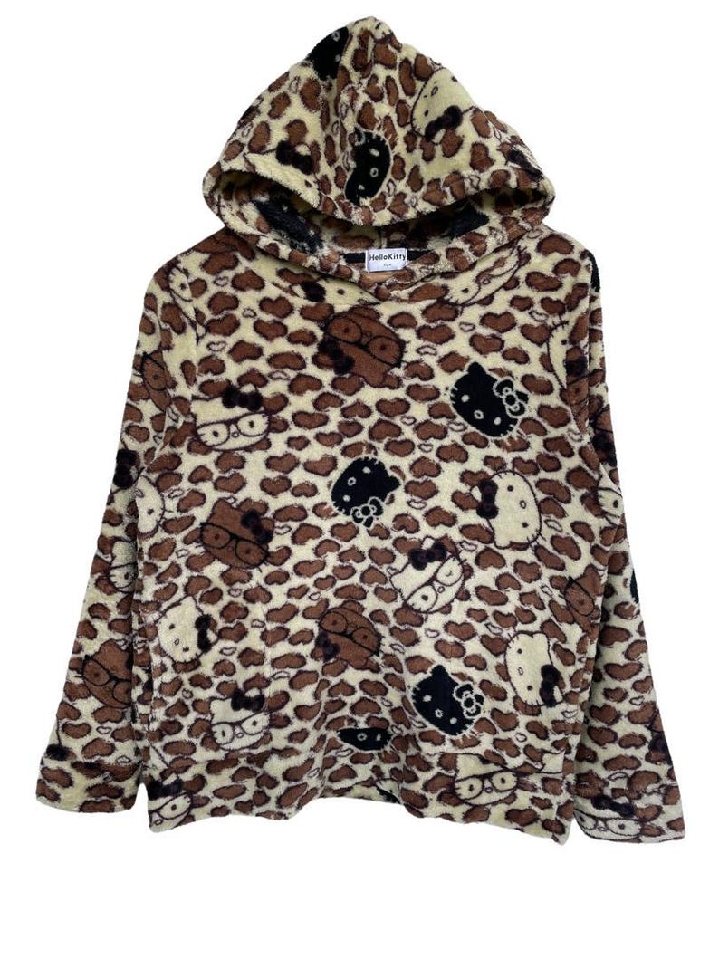 Może przedstawiać: Bluza z kapturem z wzorem w cętki i postaciami Hello Kitty. Bluza wykonana jest z miękkiego, pluszowego materiału z kapturem ze sznurkiem. Projekt zawiera brązowe, czarne i białe twarze Hello Kitty.