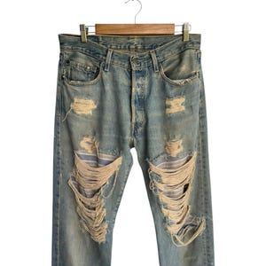 May include: Light blue denim jeans with distressed details, including multiple rips and tears across the legs. The jeans are hanging on a wooden hanger against a white background. The jeans have a button fly.