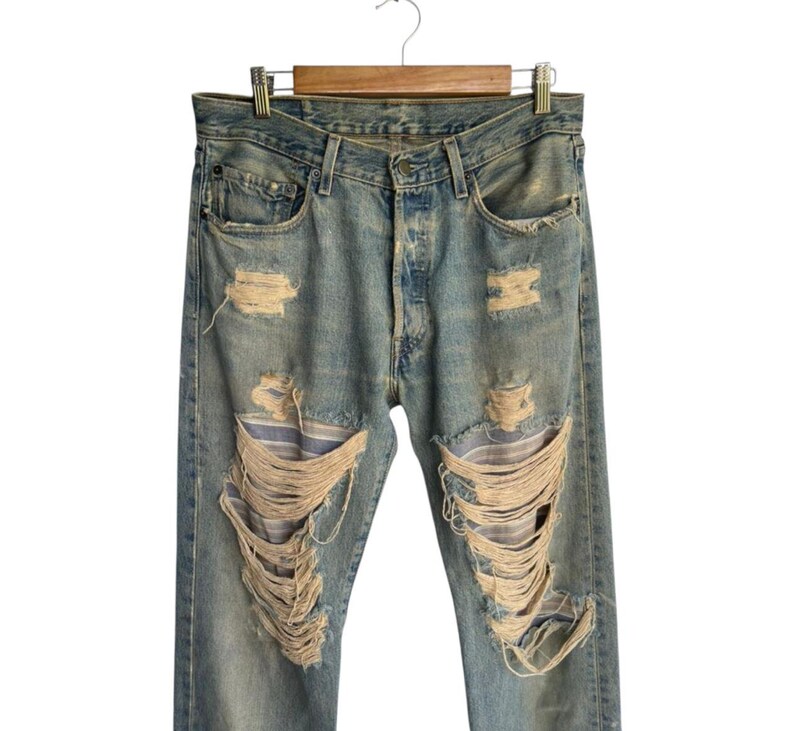 May include: Light blue denim jeans with distressed details, including multiple rips and tears across the legs. The jeans are hanging on a wooden hanger against a white background. The jeans have a button fly.