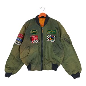 May include: Olive green bomber jacket with multiple embroidered patches. Patches include racing team logos, the text "Bugatti", and "Shoei". The jacket has a black collar, cuffs, and waistband, and a zipper closure.
