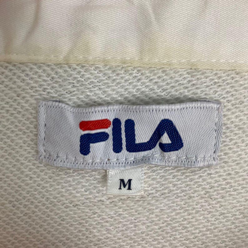 May include: A white fabric garment featuring a rectangular clothing label with the Fila logo in red and blue. Below the main label, a smaller white tag displays the letter M in black, indicating the size of the garment. The fabric has a textured knit.