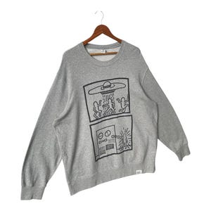 May include: A gray sweatshirt with a crew neck and long sleeves. The front features a black graphic of a UFO and figures, along with a control panel design. The sweatshirt is hanging on a wooden hanger.