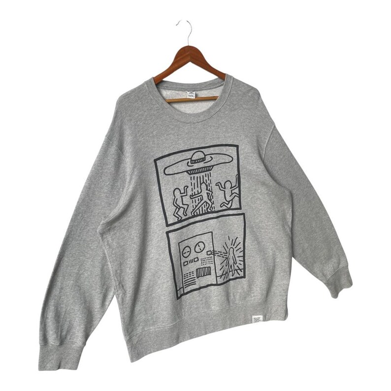 May include: A gray sweatshirt with a crew neck and long sleeves. The front features a black graphic of a UFO and figures, along with a control panel design. The sweatshirt is hanging on a wooden hanger.