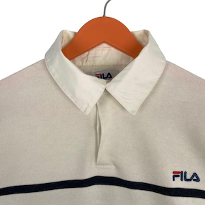 May include: A cream-coloured long-sleeve polo shirt featuring a white collar, a partial button-less placket, and a horizontal navy blue stripe across the chest. The Fila brand logo is embroidered in navy blue and red on the upper right chest area.