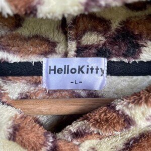Może przedstawiać: Pluszowa bluza z kapturem z brązowym, kremowym i czarnym zwierzęcym nadrukiem. Etykieta głosi "HelloKitty -L-". Odzież wykonana jest z miękkiego, puszystego materiału i prawdopodobnie jest wygodnym elementem garderoby.