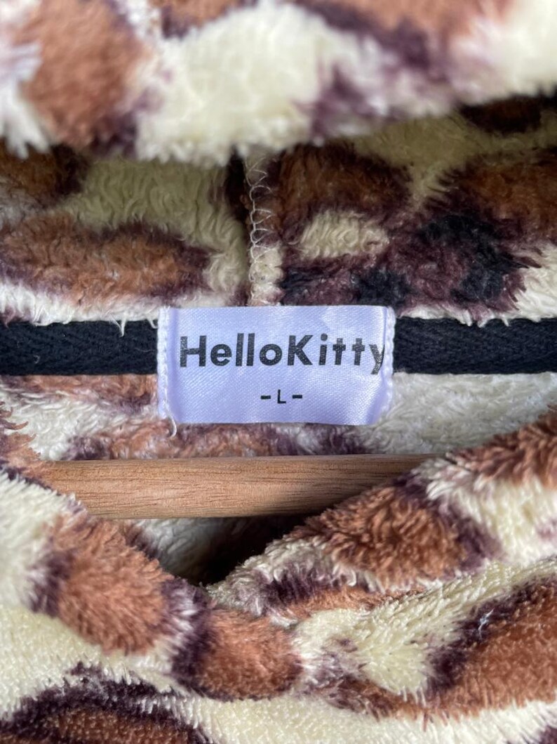 Może przedstawiać: Pluszowa bluza z kapturem z brązowym, kremowym i czarnym zwierzęcym nadrukiem. Etykieta głosi "HelloKitty -L-". Odzież wykonana jest z miękkiego, puszystego materiału i prawdopodobnie jest wygodnym elementem garderoby.