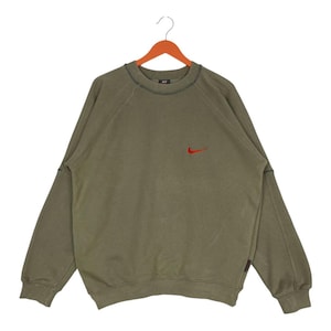 May include: Olive green sweatshirt with a small red Nike logo on the chest. The long-sleeved top has a crew neck and is hanging on a wooden hanger. The sweatshirt has a vintage, worn look.