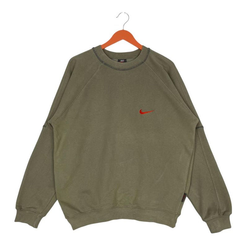 May include: Olive green sweatshirt with a small red Nike logo on the chest. The long-sleeved top has a crew neck and is hanging on a wooden hanger. The sweatshirt has a vintage, worn look.