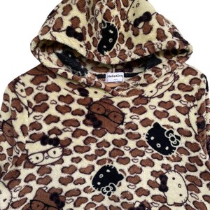 Może przedstawiać: Bluza z kapturem z nadrukiem w cętki i motywami Hello Kitty. Ubranie jest gł&oacute;wnie beżowe z brązowymi plamami i twarzami Hello Kitty w kolorze czarnym i białym. Kaptur jest podszyty czarną tkaniną.
