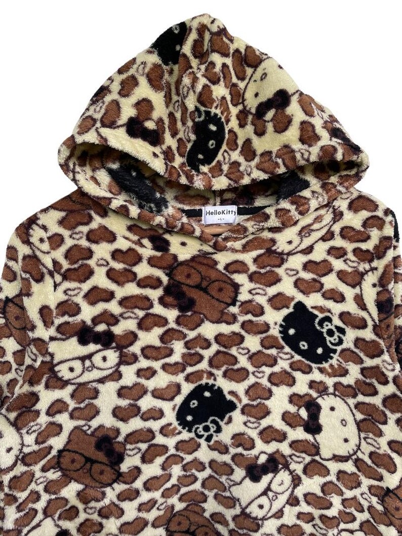 Może przedstawiać: Bluza z kapturem z nadrukiem w cętki i motywami Hello Kitty. Ubranie jest gł&oacute;wnie beżowe z brązowymi plamami i twarzami Hello Kitty w kolorze czarnym i białym. Kaptur jest podszyty czarną tkaniną.