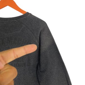 Pode incluir: Camisola cinza escuro com o texto "VENICE CA" vis&iacute;vel. A camisola tem gola redonda e mangas raglan. A imagem &eacute; um close-up do ombro e da parte superior do peito da pe&ccedil;a de vestu&aacute;rio.