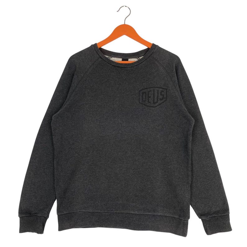 Pode incluir: Camisola cinza escuro com a palavra "DEUS" bordada no peito. A camisa de manga comprida e gola redonda est&aacute; pendurada em um cabide de madeira laranja. A camisola &eacute; feita de um material macio e confort&aacute;vel.