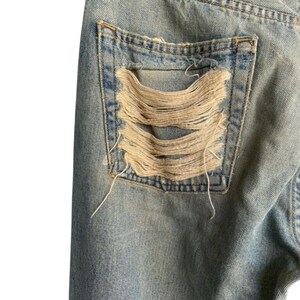 May include: Close-up of light blue denim jeans with a distressed pocket. The pocket features large, horizontal rips exposing the frayed threads. The jeans show signs of wear and tear, with a faded appearance.