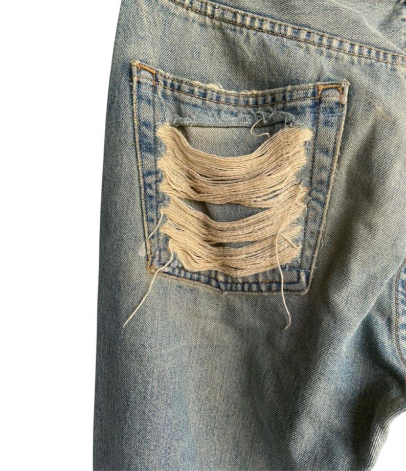 May include: Close-up of light blue denim jeans with a distressed pocket. The pocket features large, horizontal rips exposing the frayed threads. The jeans show signs of wear and tear, with a faded appearance.
