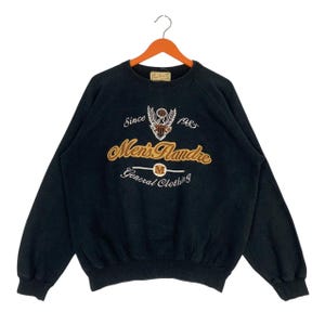 Könnte beinhalten: Ein schwarzes Langarm-Sweatshirt mit einer gestickten Grafik auf der Brust. Das Design umfasst ein Adler-Emblem, den Text "Since 1985", "Men's Flandre" und "General Clothing" mit einem kreisförmigen "M"-Logo, alles in Gold und Weiß.